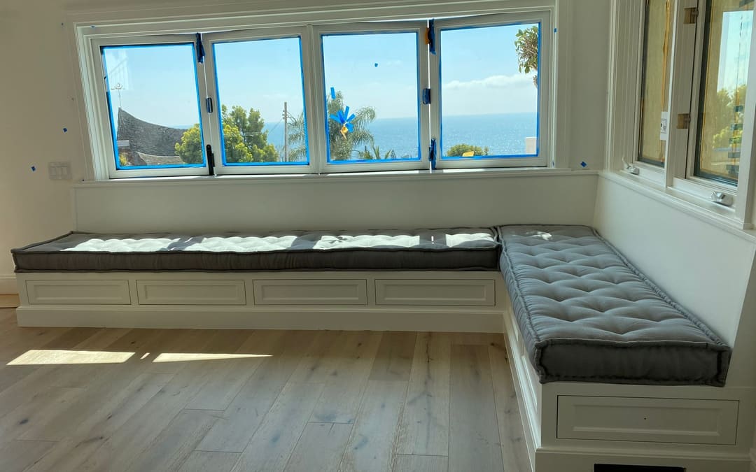 L-shaped built-in window seat with gray tufted cushions under a wall of windows facing the ocean, made by Capistrano Upholstery